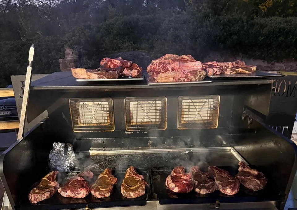 Côtes de bœuf saisies sur un grand barbecue mobile, d’autres pièces de viande en attente au-dessus, en fin de journée