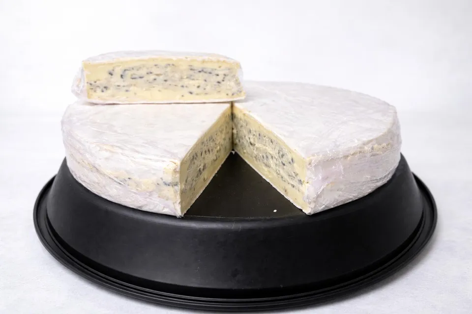 Meule de fromage à pâte persillée entamée, une part posée sur le dessus, présentée sur un plateau noir et fond blanc