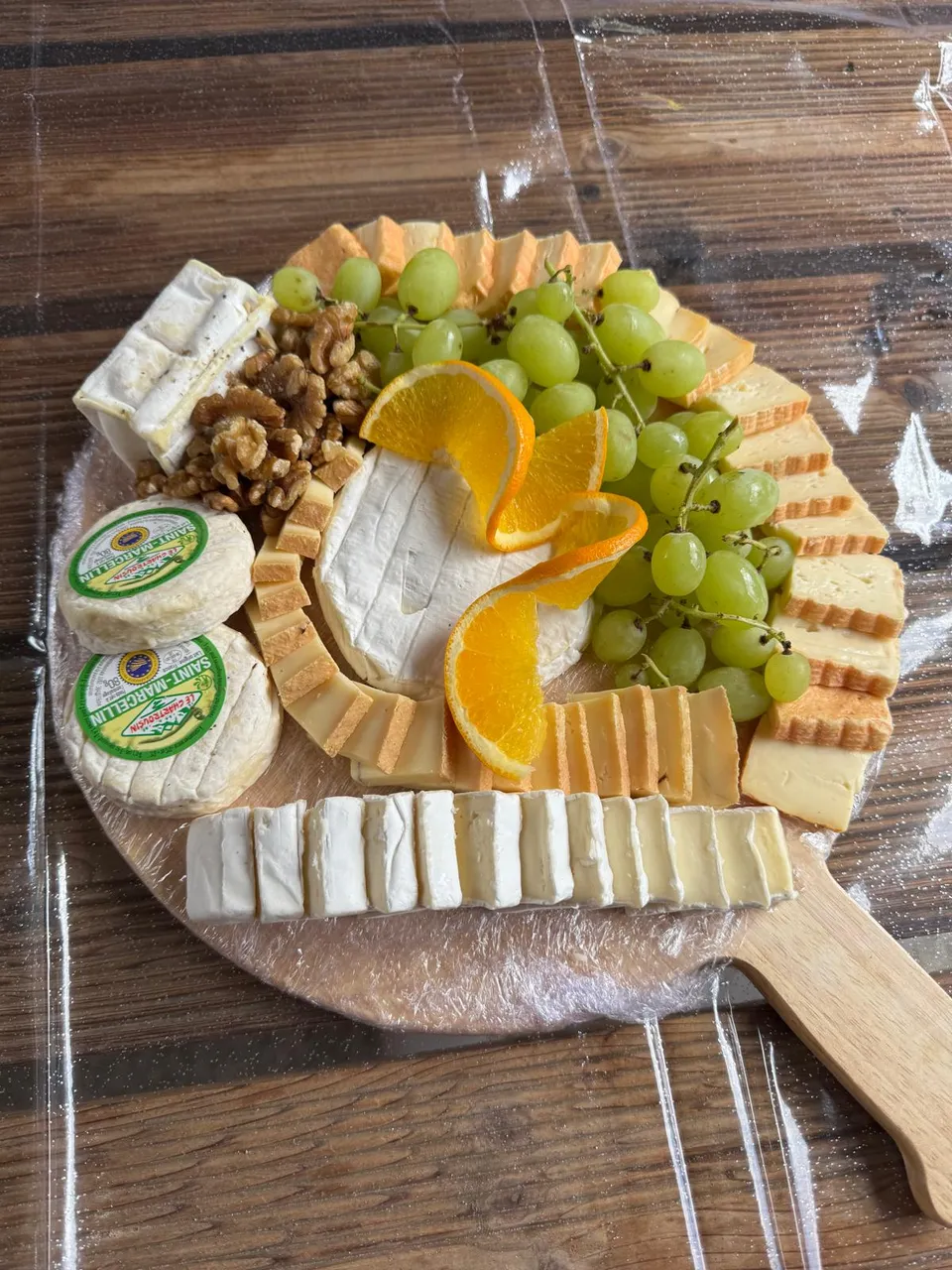 Plateau de fromages sur planche en bois : petits fromages étiquetés, brie, pâte pressée, noix, raisin blanc et oranges