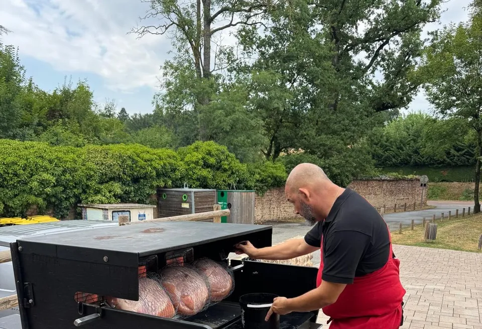 Cuisinier en tablier rouge surveillant la cuisson de jambons à la broche sur une rôtissoire mobile, en plein air