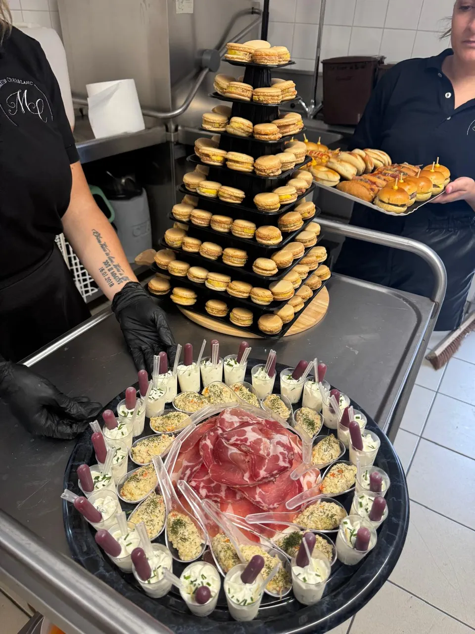 En cuisine, l’équipe dresse une pyramide de macarons, un plateau de jambon cru entouré de verrines et des mini burgers