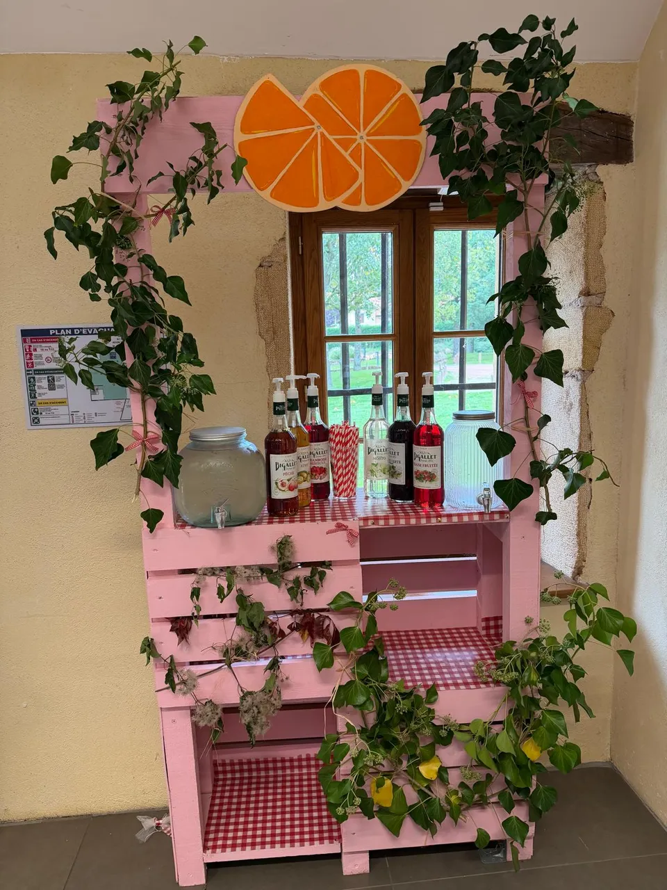 Bar à limonades en palettes roses décoré de lierre et d’oranges, avec fontaine à boisson et bouteilles de sirop