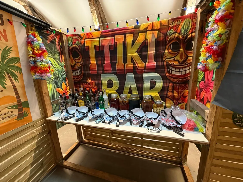 Tiki bar dressé pour une soirée : bouteilles, bocaux de fruits et verres alignés devant une toile tropicale colorée