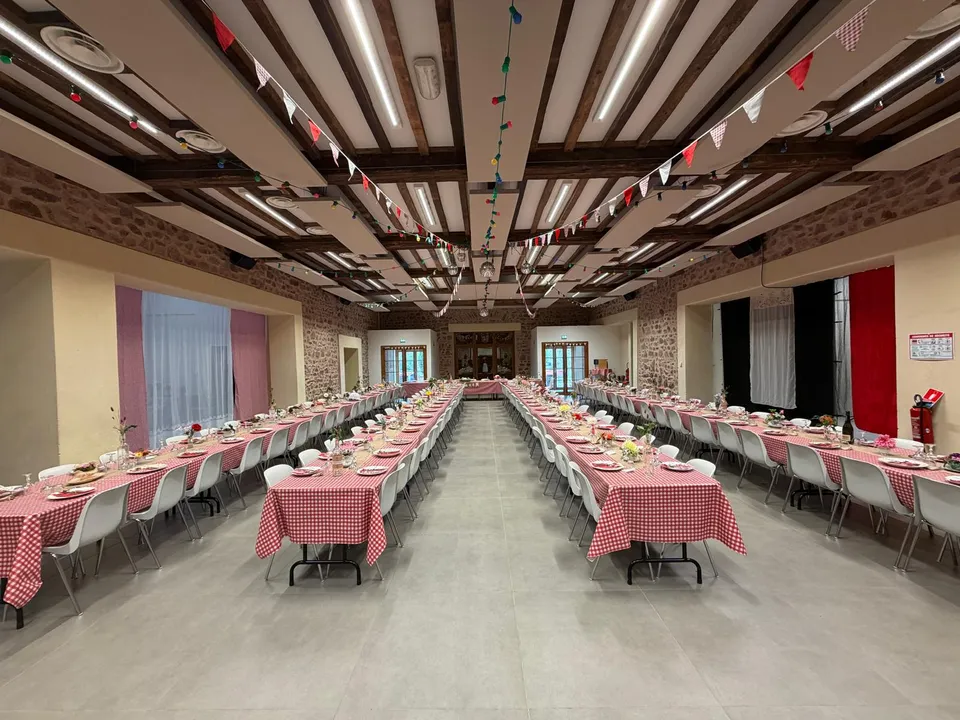 Grande salle de réception dressée : longues tables à nappes vichy rouges, chaises blanches, bouquets et fanions au plafond