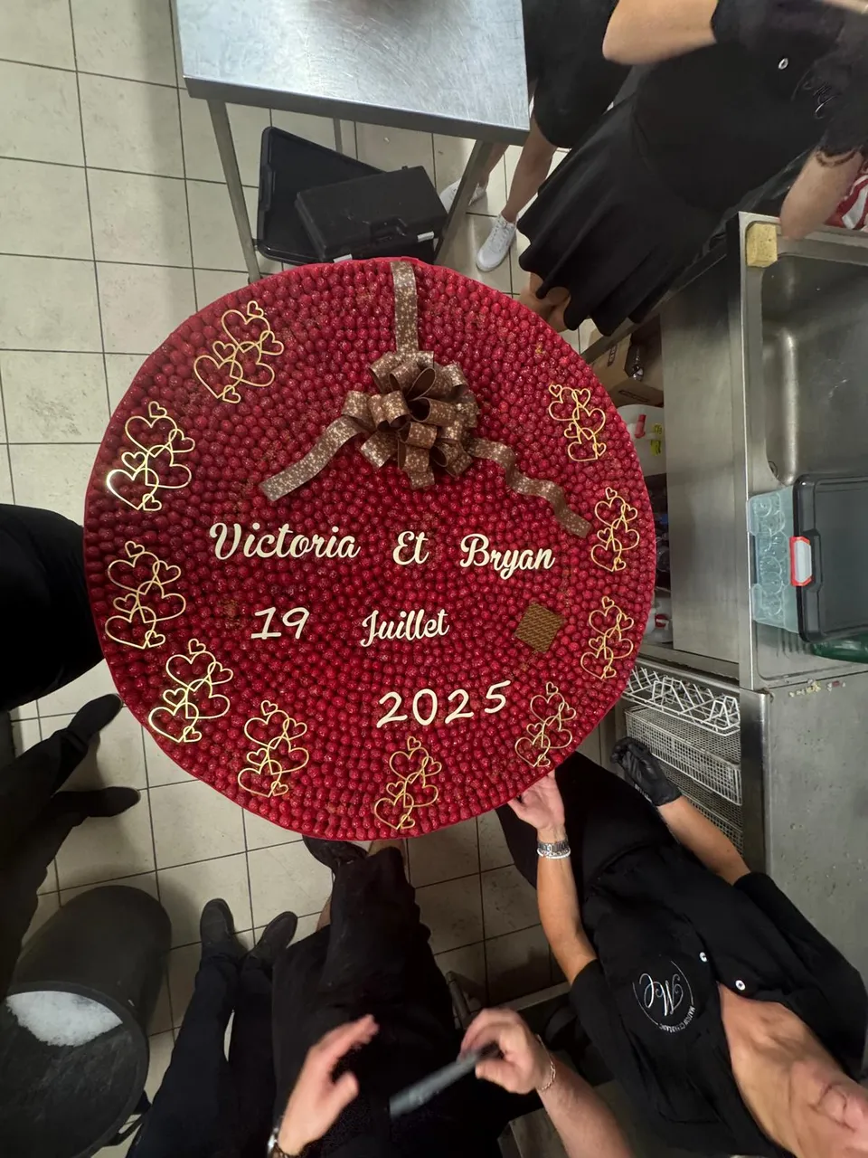 Grand gâteau de mariage rond recouvert de framboises, orné de cœurs dorés et d’un nœud en chocolat, vu de dessus