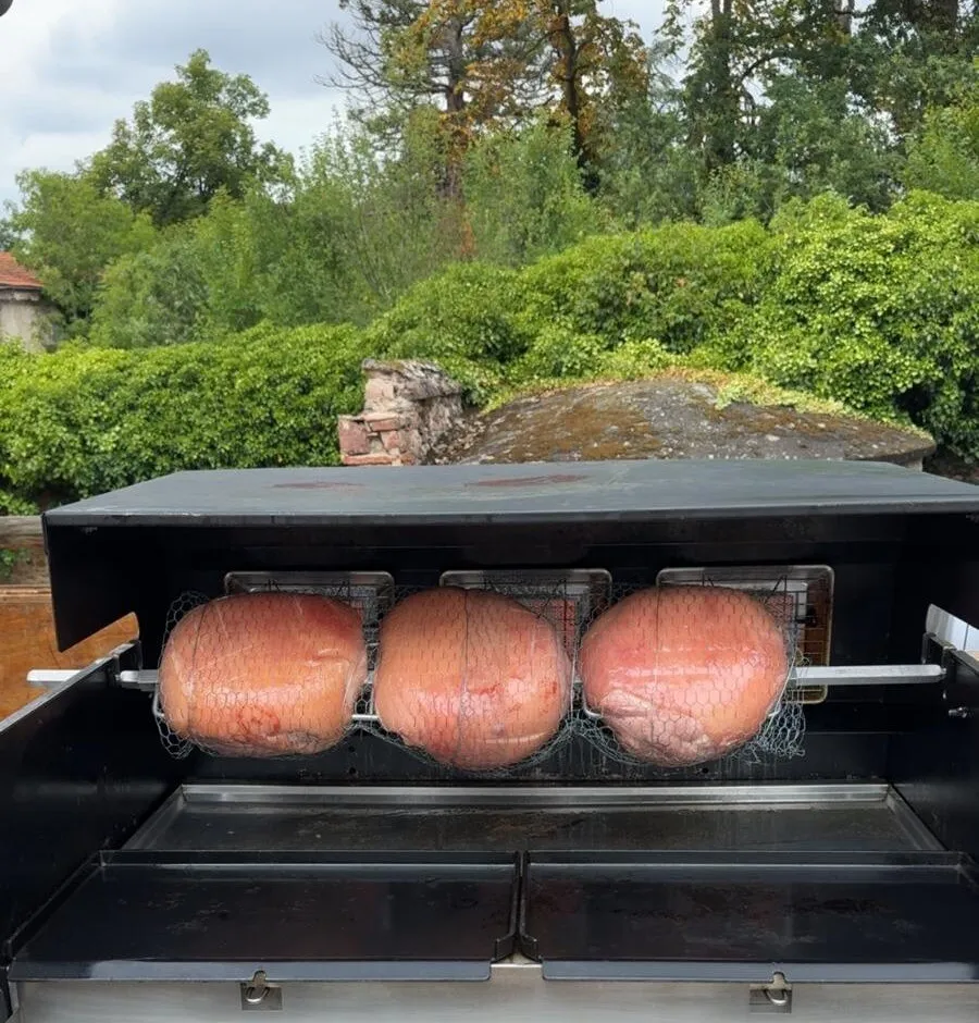 Trois jambons entiers en cuisson à la broche sur une rôtissoire mobile installée en extérieur, devant des arbres
